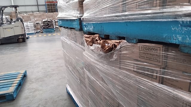 A cardboard carton on the lower pallet has been crushed and torn open directly beneath the upper pallet.