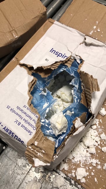 The outer cardboard carton and inner blue plastic liner are torn open, resulting in spilled product.