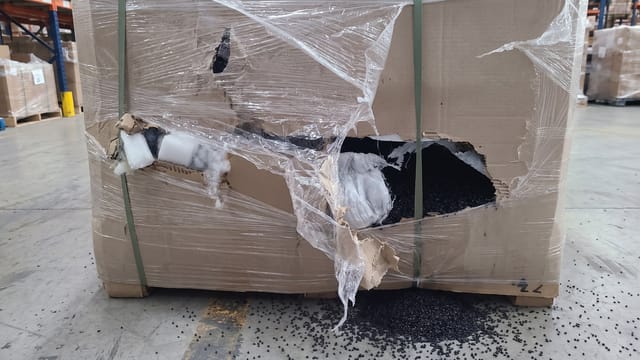 The cardboard container has a large tear on the front face, breaching containment and causing product to spill onto the floor.