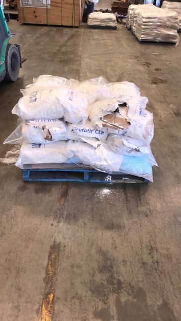 Multiple bags on the pallet are ruptured and spilling white powder onto the pallet and the floor.