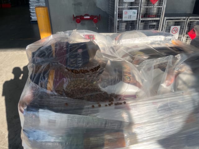 A bag of pet food on the top layer is torn, causing kibble to spill out inside the plastic wrap.