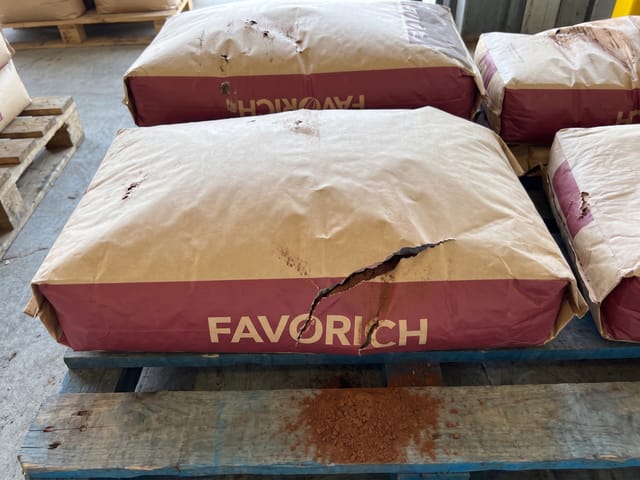 A large tear in the front paper sack has caused brown powder to spill onto the pallet, with additional minor punctures visible on other sacks.