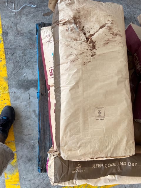 The top paper sack on the pallet is torn, exposing and spilling brown powder product.